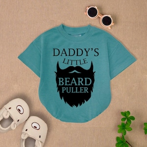May include: Teal short-sleeved baby bodysuit with the text "DADDY'S LITTLE BEARD PULLER" and a black beard graphic. The image also includes a pair of white slippers and round sunglasses.