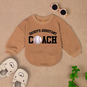 May include: Tan long-sleeve sweatshirt with a baseball graphic and the text "Daddy's Assistant Coach".