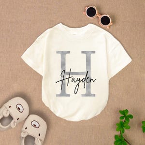 May include: White short-sleeved t-shirt with a silver letter H and the name Hayden written in cursive underneath.