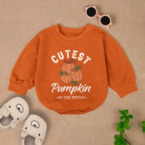 May include: Orange baby bodysuit with the text "Cutest Pumpkin" and a pumpkin graphic. The long-sleeved outfit has a round neckline and is paired with white slippers and sunglasses.