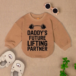May include: A brown long-sleeved sweatshirt with a black dumbbell graphic and the text "Daddy's Future Lifting Partner".