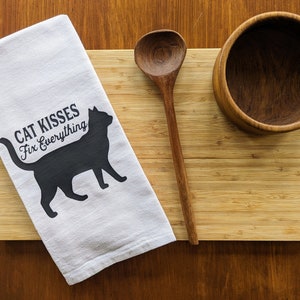 May include: White kitchen towel with a black silhouette of a cat and the text "Cat Kisses Fix Everything". The towel is lying on a wooden cutting board with a wooden spoon and bowl.