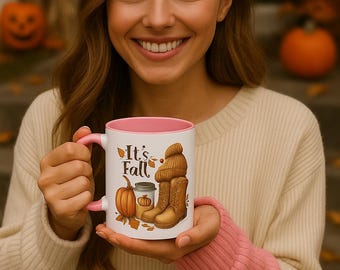 May include: White ceramic mug with a pink interior and handle, featuring the text "It's Fall" and autumn-themed illustrations of a pumpkin, boots, and a hat. The mug is held by a person wearing a cream-colored sweater.
