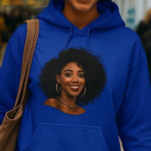 Könnte beinhalten: Ein königsblauer Kapuzenpullover mit einem Porträt einer Frau mit lockigem Haar, goldenen Ohrringen und einer Halskette. Der Kapuzenpullover hat eine Vordertasche und eine Kapuze mit Kordelzug. Die Person lächelt und hält einen Koffer.