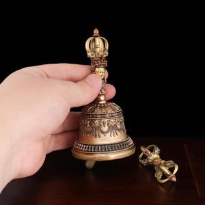 May include: A gold-colored Tibetan bell with a detailed design and a crown-shaped top. The bell is held in a hand and a matching dorje is on the table.