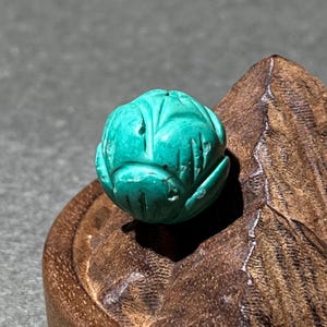 May include: A turquoise-coloured carved stone bead, shaped like a flower bud. The bead features detailed carvings and rests on a wooden base. The turquoise colour is vibrant and the carving is intricate.