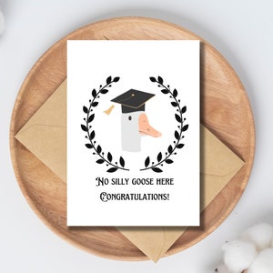 May include: A white card with a black and white illustration of a goose wearing a graduation cap. The text on the card reads "No silly goose here Congratulations!"