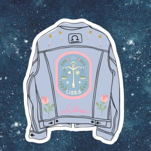 May include: A light blue denim jacket with a pink and white Libra zodiac sign patch on the back. The patch features a scale, stars, and flowers. The jacket also has two pink roses embroidered on the bottom.