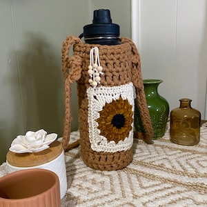 May include: A brown crocheted water bottle holder with a sunflower design. The holder has a strap for carrying and wooden beads.