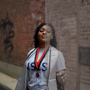 May include: A woman wearing a gray graduation gown and a gray shirt with the word "SLS" printed on it. She is wearing a red and black graduation cord around her neck and a medal. She has long black braids and is smiling.
