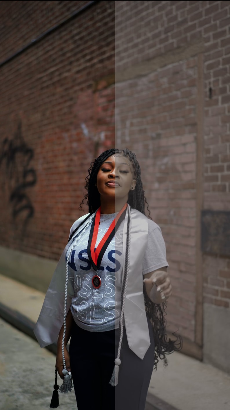 May include: A woman wearing a gray graduation gown and a gray shirt with the word "SLS" printed on it. She is wearing a red and black graduation cord around her neck and a medal. She has long black braids and is smiling.