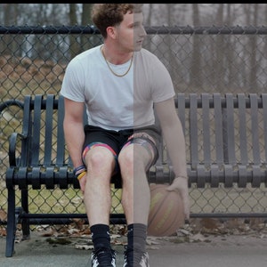 May include: A young man wearing a white t-shirt, black shorts, and black socks sits on a black metal bench. He is holding a basketball in his hands.