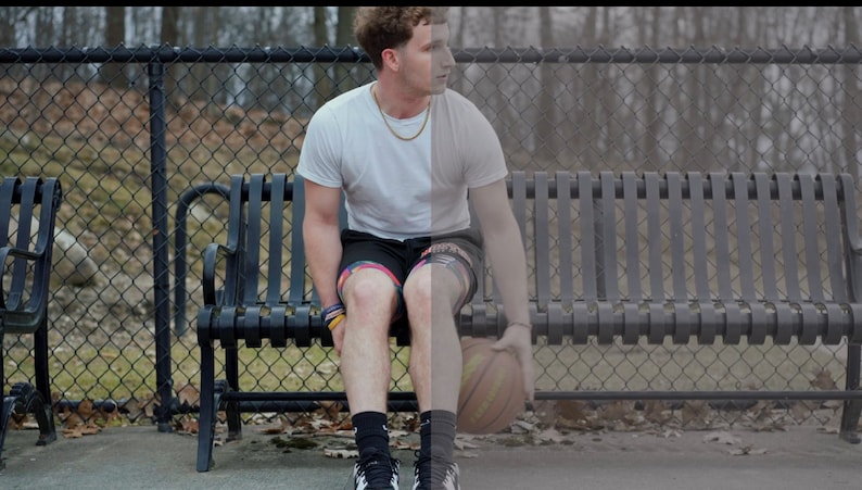 May include: A young man wearing a white t-shirt, black shorts, and black socks sits on a black metal bench. He is holding a basketball in his hands.
