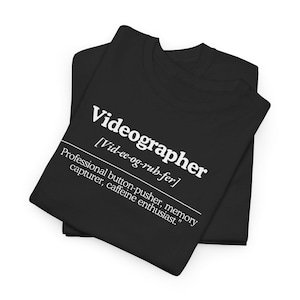 May include: Black t-shirt with white text that reads "Videographer [Vid-ee-og-rub-fer] Professional button-pusher, memory capturer, caffeine enthusiast."