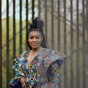 May include: A woman wearing a short, colorful dress with a geometric pattern. The dress has ruffled sleeves and a plunging neckline. She is standing in front of a metal fence.