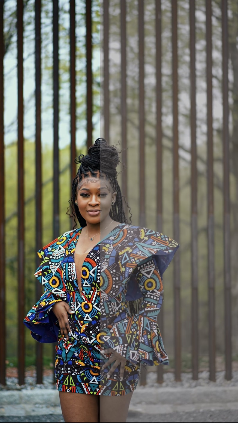 May include: A woman wearing a short, colorful dress with a geometric pattern. The dress has ruffled sleeves and a plunging neckline. She is standing in front of a metal fence.