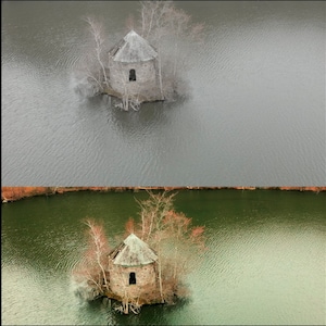 May include: An aerial view of a small stone building with a green roof on a small island in a lake. The water is calm and the sky is gray.