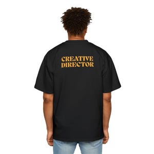 以下が含まれることがあります： 背面にオレンジ色の「CREATIVE DIRECTOR」の文字がプリントされた黒いTシャツです。