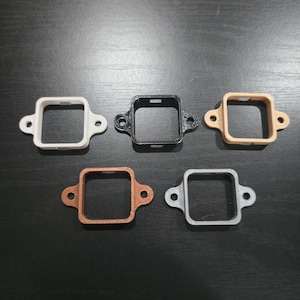 May include: Five square-shaped watch case protectors in various colors: white, black, tan, copper, and silver. Each case has a square opening and two circular holes on either side.