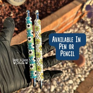 May include: Two beaded pens, one with blue, green, and black beads, and the other with white and green beads. The pens are held in a gloved hand. The text "AVAILABLE IN PEN OR PENCIL" is on a blue background.