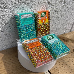 May include: Four Tic Tac mint containers decorated with colorful rhinestones. The containers are arranged in a row on a white surface. The containers are labeled "tic tac" and "wintergreen".