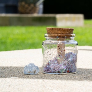 Op de afbeelding: Een glazen pot met een kurken deksel gevuld met iriserende glitter. De pot staat op een lichtgrijze ondergrond met een klein, helder, steenachtig voorwerp ernaast.