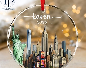 May include: A circular, faceted glass ornament featuring a printed New York City skyline with the Statue of Liberty, the text "karen" in white script, and the year "2026" below it. A red ribbon is attached to the top for hanging purposes.