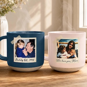 May include: A dark blue ceramic mug featuring a photo of a child and adult with the text Daddy Est. 1998 and a light pink ceramic mug featuring a photo of an adult and two children with the text We love you, Mom, both with speckled bases.