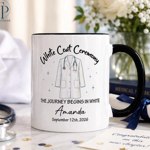 May include: A white ceramic mug with a black handle featuring a graphic of a white lab coat and a stethoscope. The text reads White Coat Ceremony, The Journey Begins In White, Amanda, and September 12th, 2026, surrounded by small blue star icons.