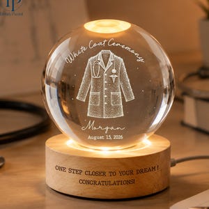 May include: A clear glass sphere lamp on a round wooden base. The sphere features an etched white coat, a stethoscope, the text White Coat Ceremony, the name Morgan, and August 15, 2026. The base is engraved with ONE STEP CLOSER TO YOUR DREAM! CONGRATULATIONS!
