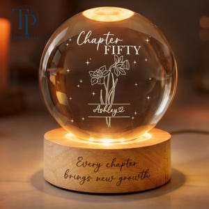 May include: A glowing glass sphere with the words "Chapter FIFTY" and "Ashley" engraved on it, along with a floral design. The sphere sits on a wooden base with the inscription "Every chapter brings new growth."