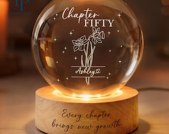 May include: A glowing glass sphere with the words "Chapter FIFTY" and "Ashley" engraved on it, along with a floral design. The sphere sits on a wooden base with the inscription "Every chapter brings new growth."