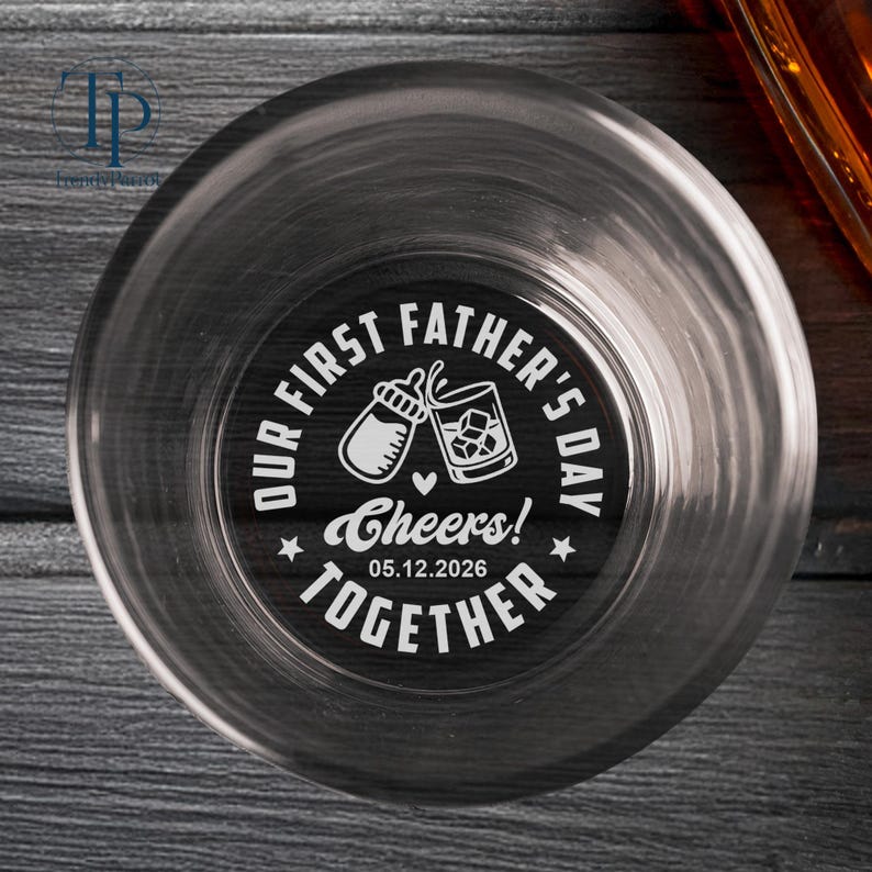 May include: Clear glass tumbler with a black and white design. The design includes the text "OUR FIRST FATHER'S DAY" and "Cheers! Together 05.12.2026" around a baby bottle and a whiskey glass with ice cubes.