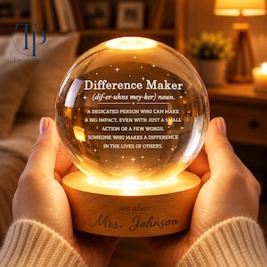 May include: A lit glass sphere with the definition of "Difference Maker" inscribed within, resting on a wooden base. The base is engraved with the name "Mrs. Johnson". The sphere emits a warm light, creating a cosy atmosphere.