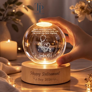May include: A clear glass sphere lamp on a round wooden base. The sphere features an etched stethoscope, floral accents, and the text: Never underestimate the difference you have made and the lives you have touched, Sophia, RN. The base reads: Happy Retirement Sep 2026.