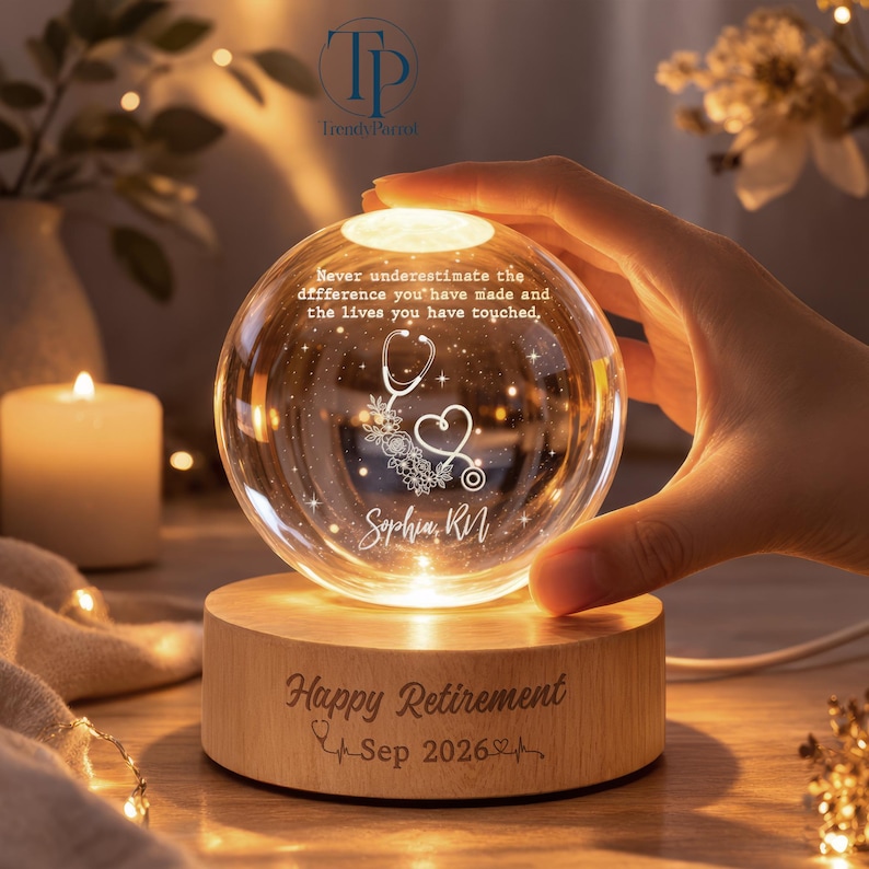 May include: A clear glass sphere lamp on a round wooden base. The sphere features an etched stethoscope, floral accents, and the text: Never underestimate the difference you have made and the lives you have touched, Sophia, RN. The base reads: Happy Retirement Sep 2026.