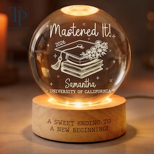 May include: A graduation-themed crystal ball with the text "Mastered It!" and the year 2026. The crystal ball features a graduation cap, books, and the name "Samantha" with "UNIVERSITY OF CALIFORNIA". It sits on a wooden base with the inscription "A SWEET ENDING TO A NEW BEGINNING!"