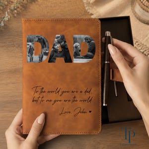 K&ouml;nnte beinhalten: Ein braunes Lederjournal mit dem ausgeschnittenen Wort "DAD", das Schwarz-Wei&szlig;-Fotos zeigt. Das Journal enth&auml;lt einen Stift und die Inschrift "To the world you are a dad, but to me you are the world. Love, John."