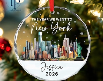May include: A circular clear acrylic ornament featuring a colorful New York City skyline illustration, the Statue of Liberty, and white snowflakes. The text reads THE YEAR WE WENT TO New York, Jessica, and 2026. It hangs from a red ribbon.