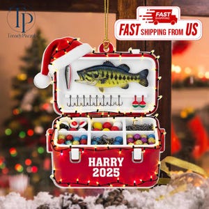 May include: A red tackle box ornament featuring a Santa hat on top and string lights wrapped around the frame. The interior displays a bass fish, hooks, and colorful fishing lures. The front is printed with the text HARRY 2025 in white letters.