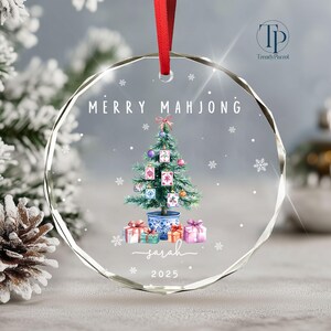 May include: A clear, round Christmas ornament with a red ribbon. The ornament features a watercolor illustration of a Christmas tree decorated with Mahjong tiles, gifts, and the text "MERRY MAHJONG". Below the tree is the name "Sarah" and the year "2025".