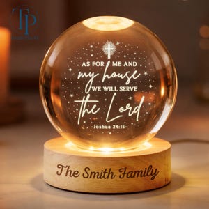 May include: A lit crystal ball night light with the text "AS FOR ME AND my house WE WILL SERVE the Lord - Joshua 24:15-". It rests on a wooden base engraved with "The Smith Family". The light is a decorative item.