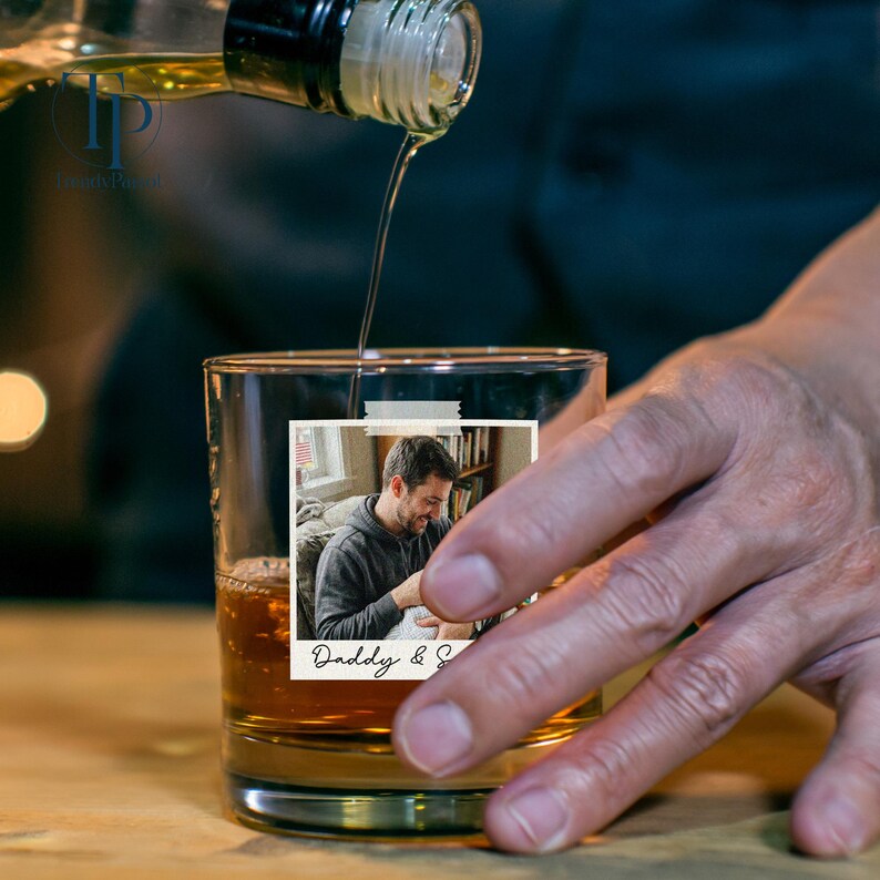 May include: A clear glass tumbler with a photo of a man taped to the front, filled with amber liquid as liquor is poured in. The photo has the text "Daddy & S".