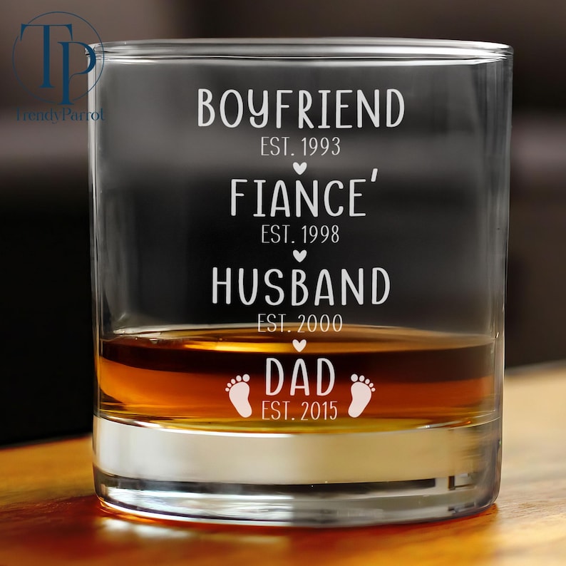 May include: A clear glass tumbler with the text "Boyfriend Est. 1993", "Fiance' Est. 1998", "Husband Est. 2000", and "Dad Est. 2015" etched on the side. The glass is filled with amber liquid.