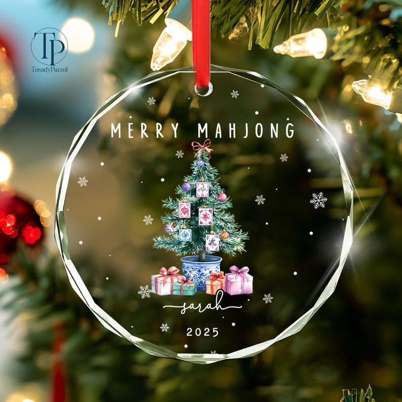 May include: A clear glass Christmas ornament with the text "MERRY MAHJONG" and "sarah 2025". The ornament features a Christmas tree decorated with Mahjong tiles, gifts, and snowflakes. A red ribbon is attached for hanging.