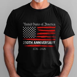 May include: Black t-shirt featuring a distressed American flag design in red and white. The text "United States of America" arches above the flag, with "250TH ANNIVERSARY" below. The years "1776-2026" are printed at the bottom.