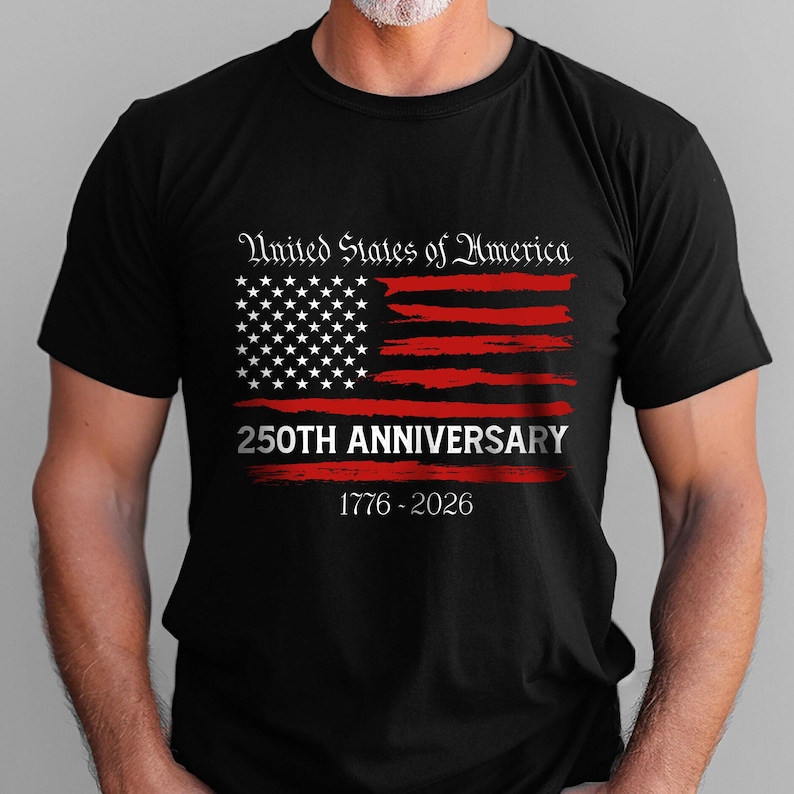 May include: Black t-shirt featuring a distressed American flag design in red and white. The text "United States of America" arches above the flag, with "250TH ANNIVERSARY" below. The years "1776-2026" are printed at the bottom.