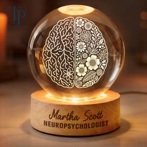 May include: A clear glass sphere lamp featuring an etched design of a brain with one side showing neural pathways and the other side showing floral patterns. The sphere sits on a round wooden base engraved with the text Martha Scott NEUROPSYCHOLOGIST.