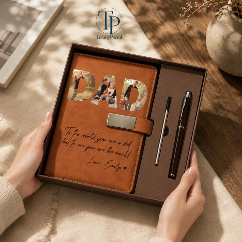 K&ouml;nnte beinhalten: Ein braunes Lederjournal mit dem Wort "DAD" ausgeschnitten, um Familienfotos freizugeben. Das Journal enth&auml;lt einen Stift und einen Bleistift sowie die Inschrift "To the world you are a dad, but to me you are the world. Love, Emily."