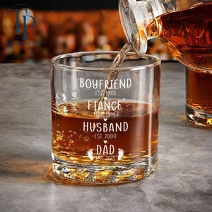 May include: A clear glass with the text "Boyfriend Est. 1993, Fianc&eacute; Est. 1998, Husband Est. 2000, Dad Est. 2015" and a pair of footprints. The glass is filled with amber liquid.